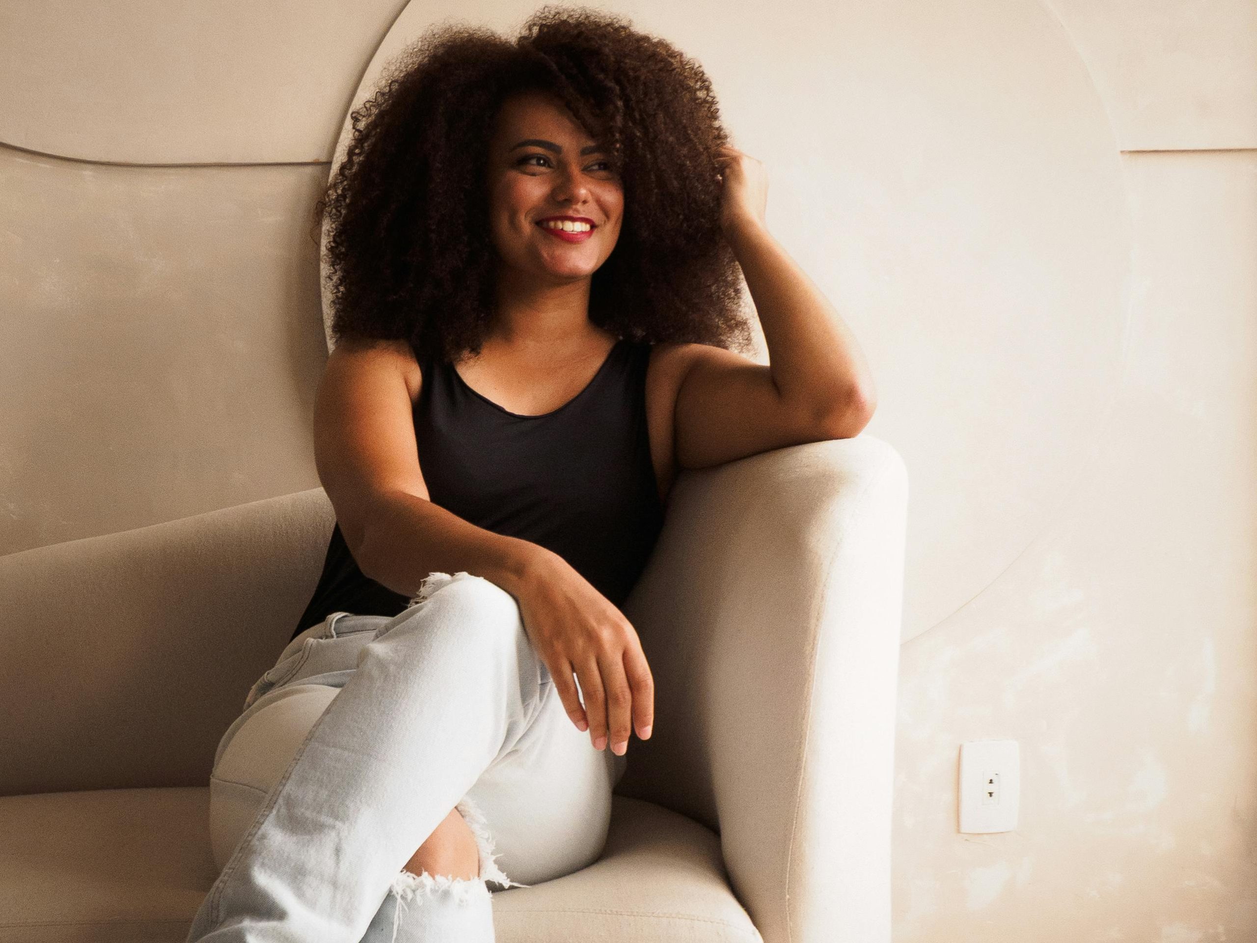 A joyful woman with curly hair relaxing indoors on a comfortable armchair, exuding warmth and contentment.